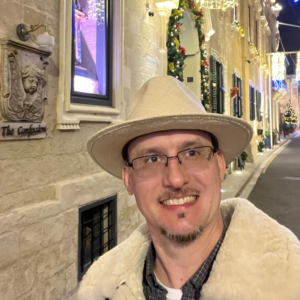 A person wearing glasses and a beige hat smiles while taking a selfie on a festive, decorated street. The stone buildings are adorned with Christmas lights and garlands. In the background, a Christmas tree is visible, adding to the holiday atmosphere.